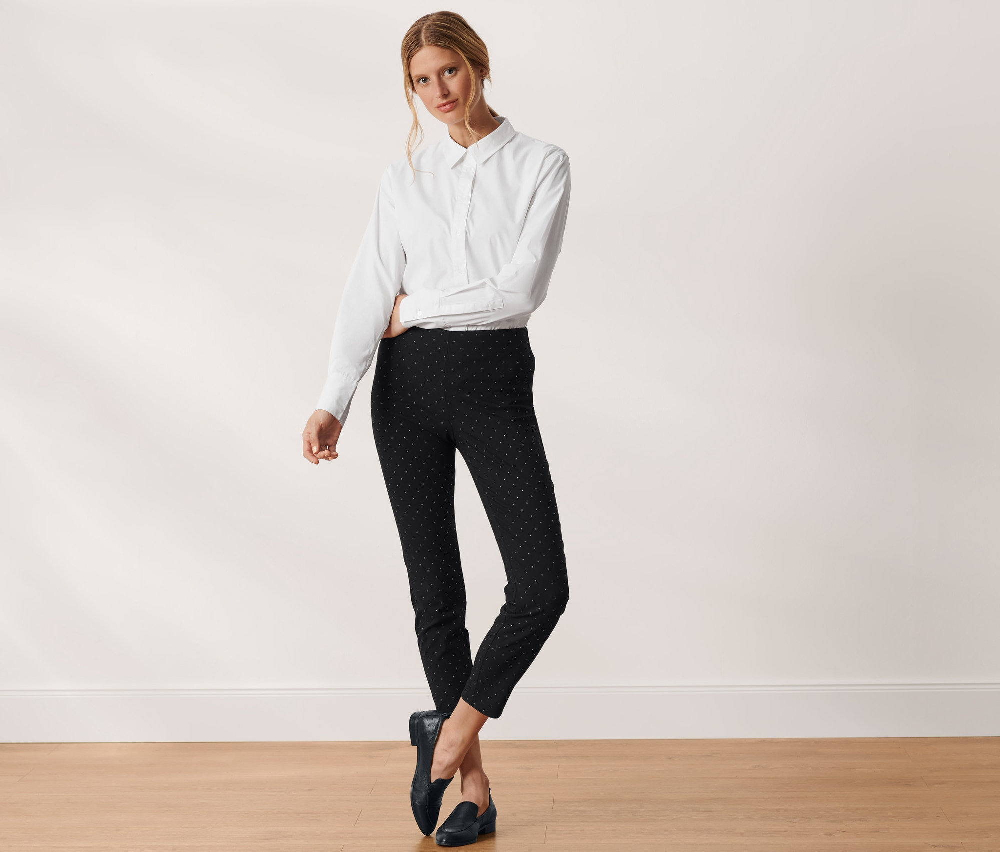 Žena pózuje v bielej košeli a čiernych bodkovaných elastických nohaviciach.