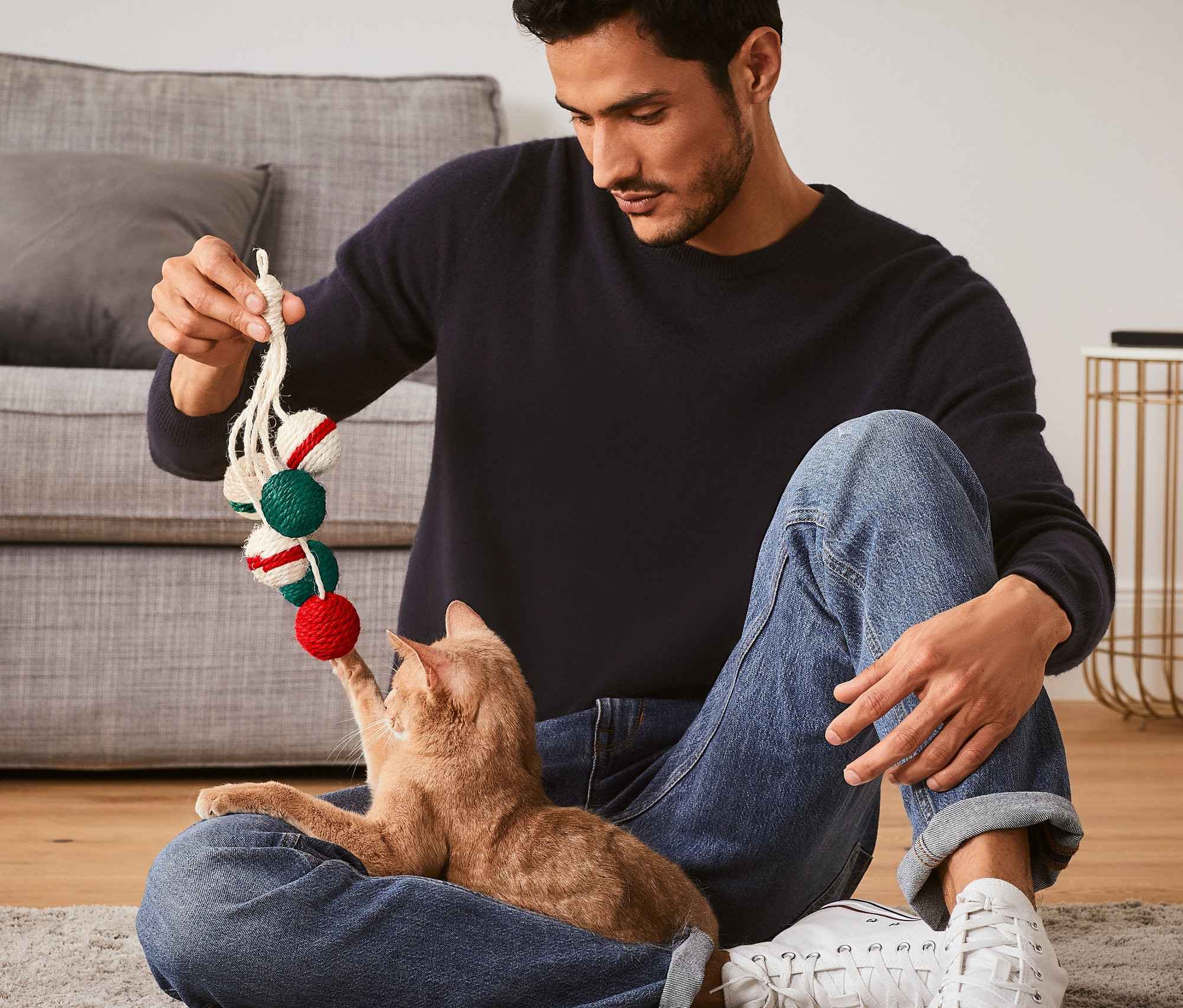 Muž sedí na podlahe a hrá sa s mačkou s hračkou pre mačky, hrkálka.