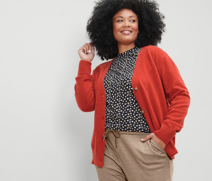 Žena pózuje v kardigáne s výstrihom do V, béžových teplákových nohaviciach a tričku s dlhými rukávmi a celoplošnou potlačou.