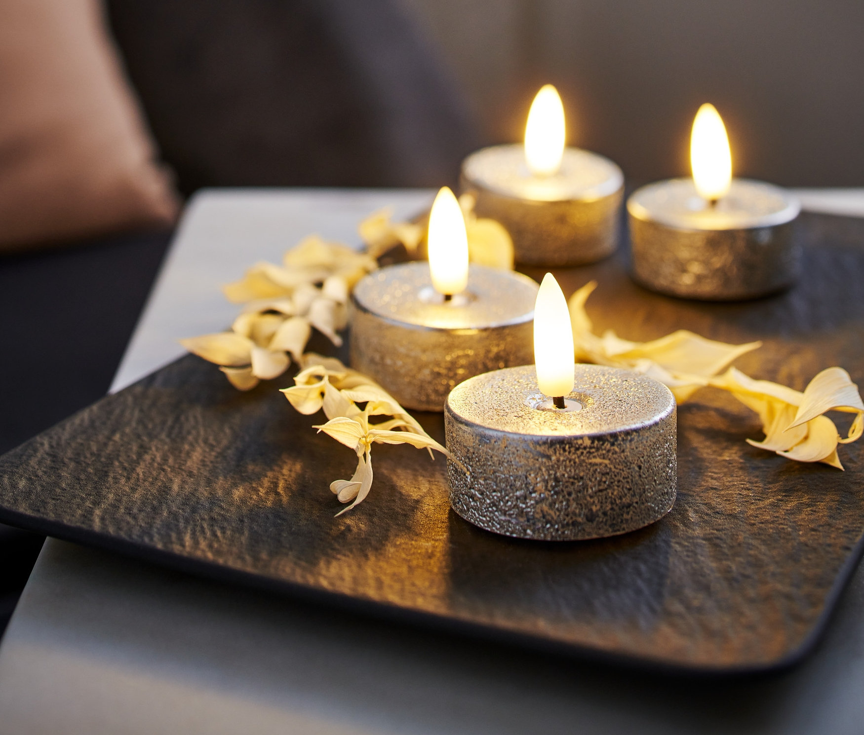 Štyri strieborné LED čajové sviečky z pravého vosku na čiernej podložke, zdobené suchými kvetmi.