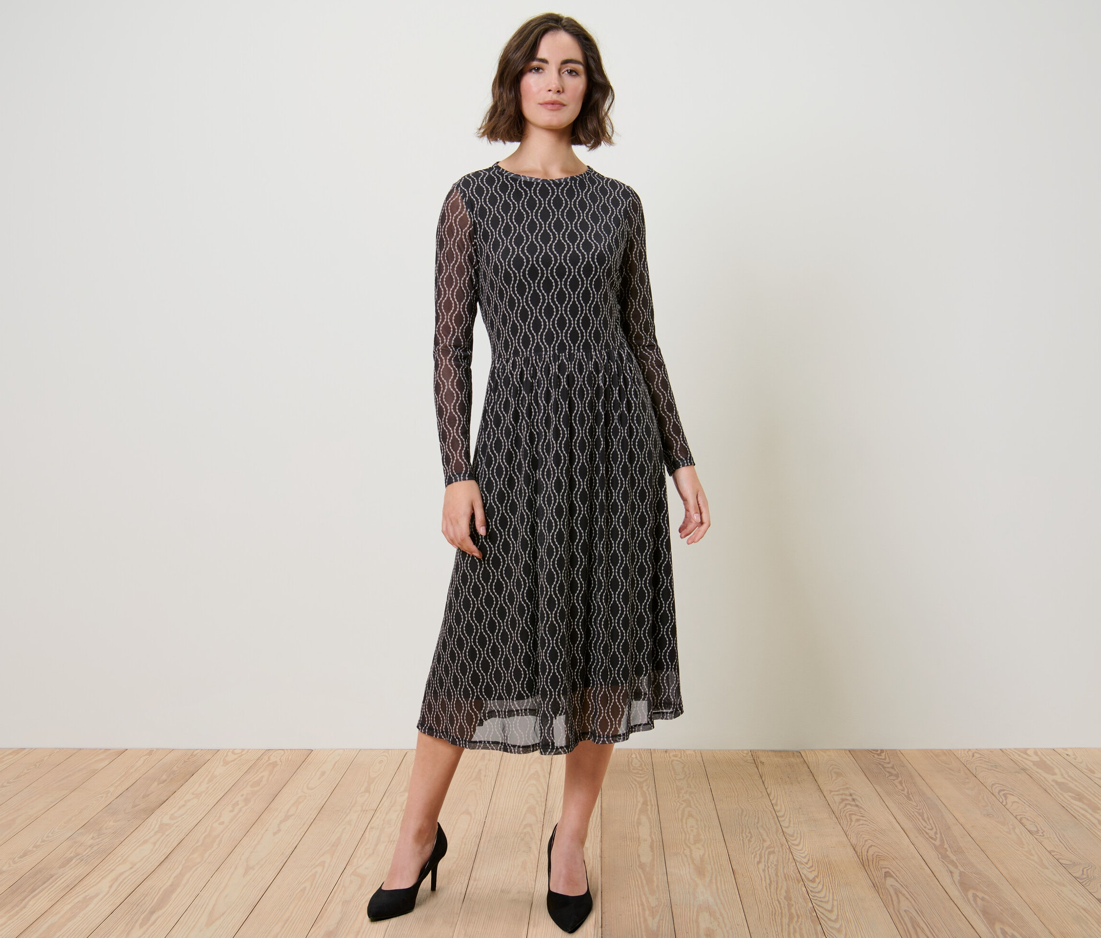 Žena pózuje v šatách zo sieťoviny s potlačou a čiernych lodičkách.