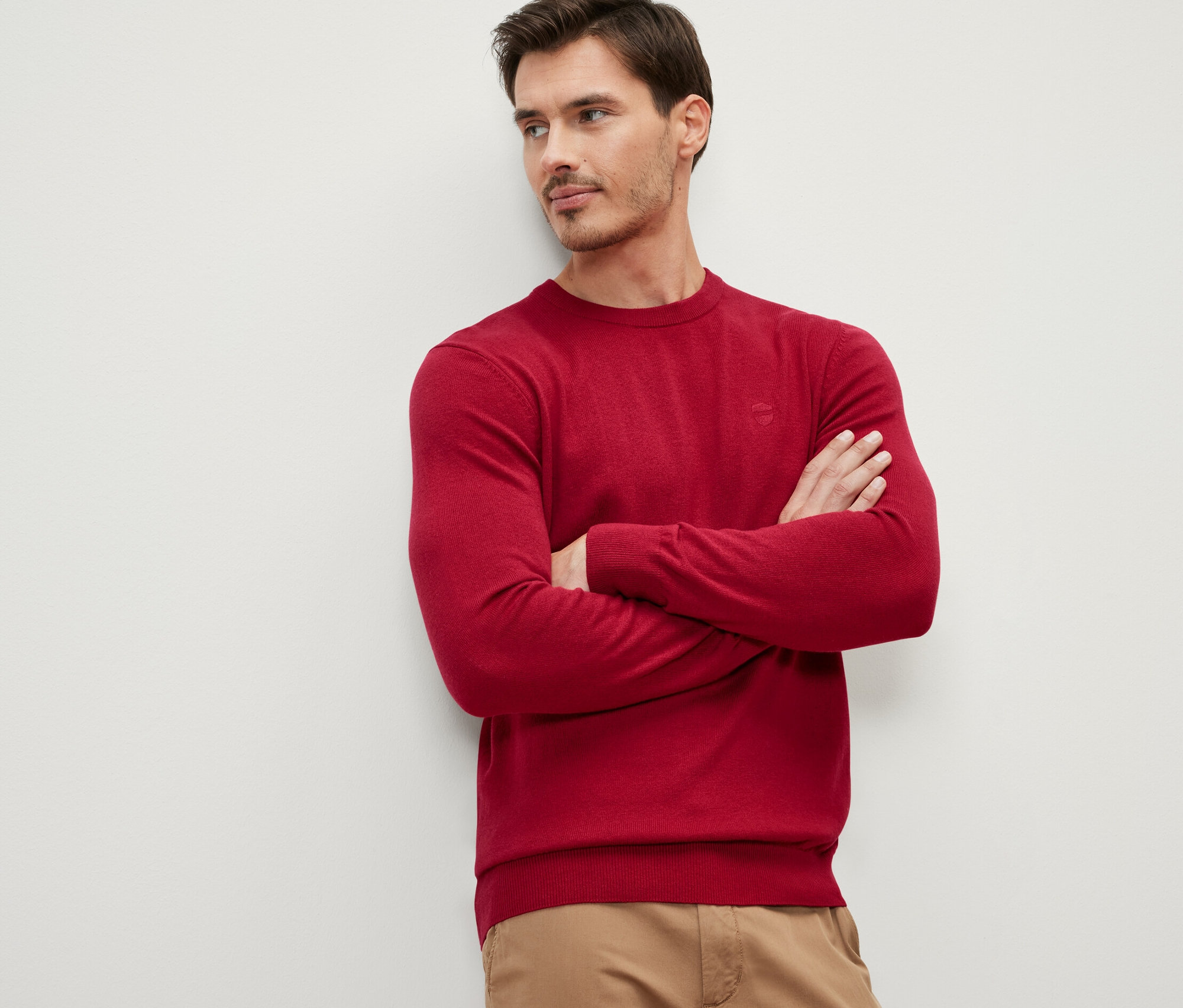 Muž so skríženými rukami na hrudi má na sebe červený pulóver s okrúhlym výstrihom.