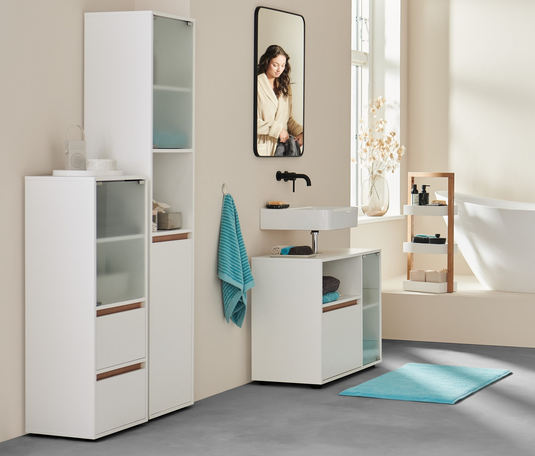 V kúpeľni stojí žena v župane pri vani. V popredí komoda, kúpeľňa, mimoriadne veľká osuška antracitovej farby a 2 ks akvamarínové uteráky.