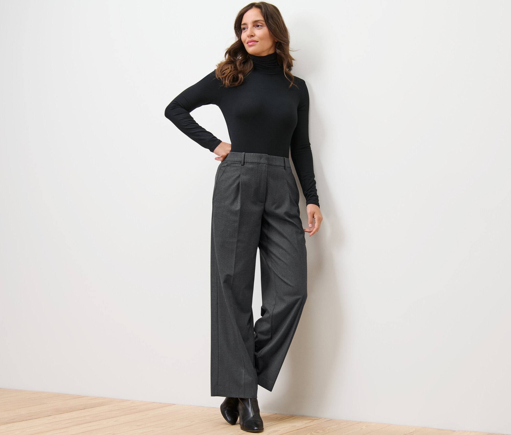 Žena pózuje opretá o stenu v čiernom roláku a pohodlných tkaných nohaviciach.