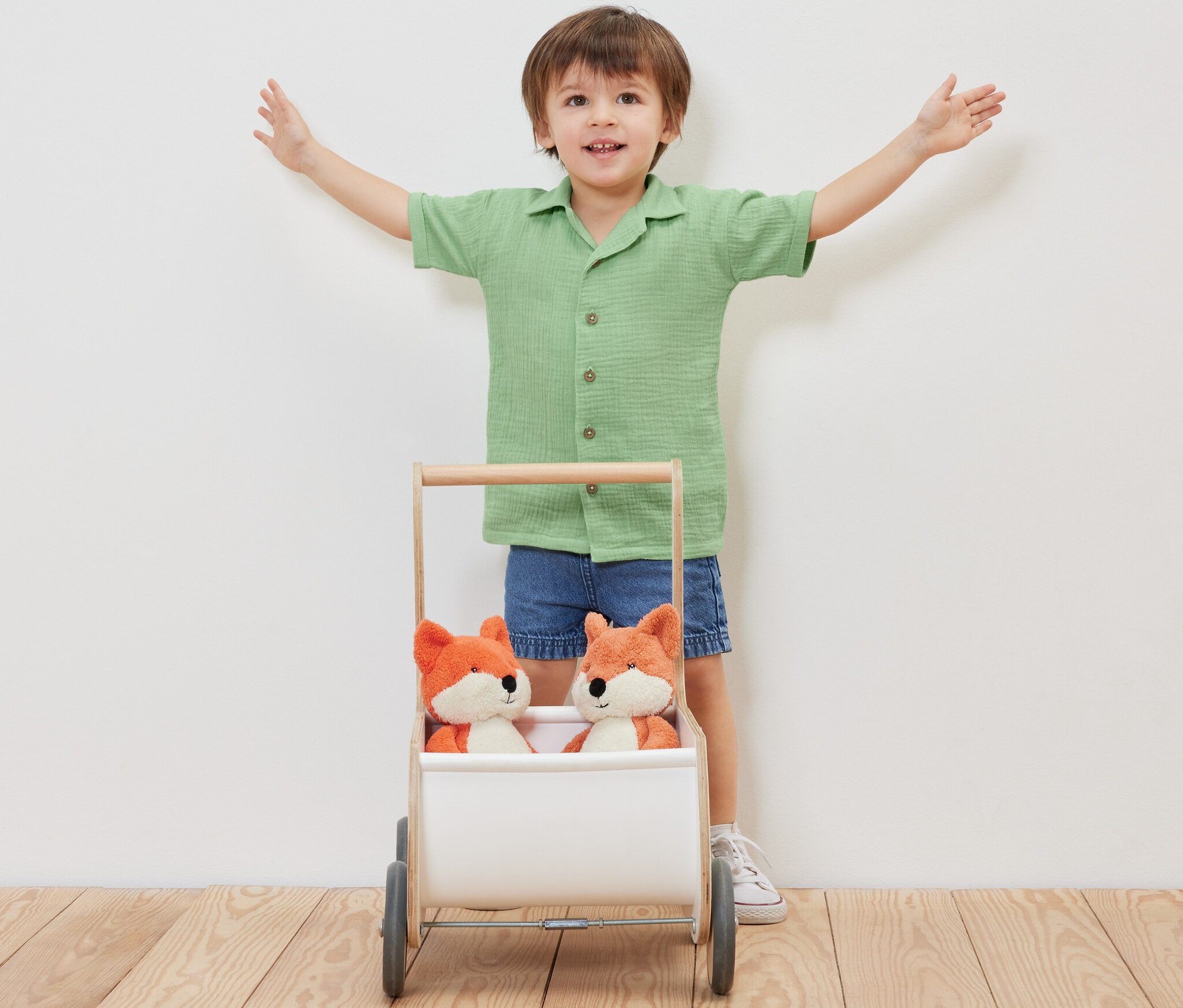 Chlapec stojí s rozpaženými rukami za vozíkom s dvoma plyšovými líškami a má na sebe detskú mušelínovú košeľu s golierom.