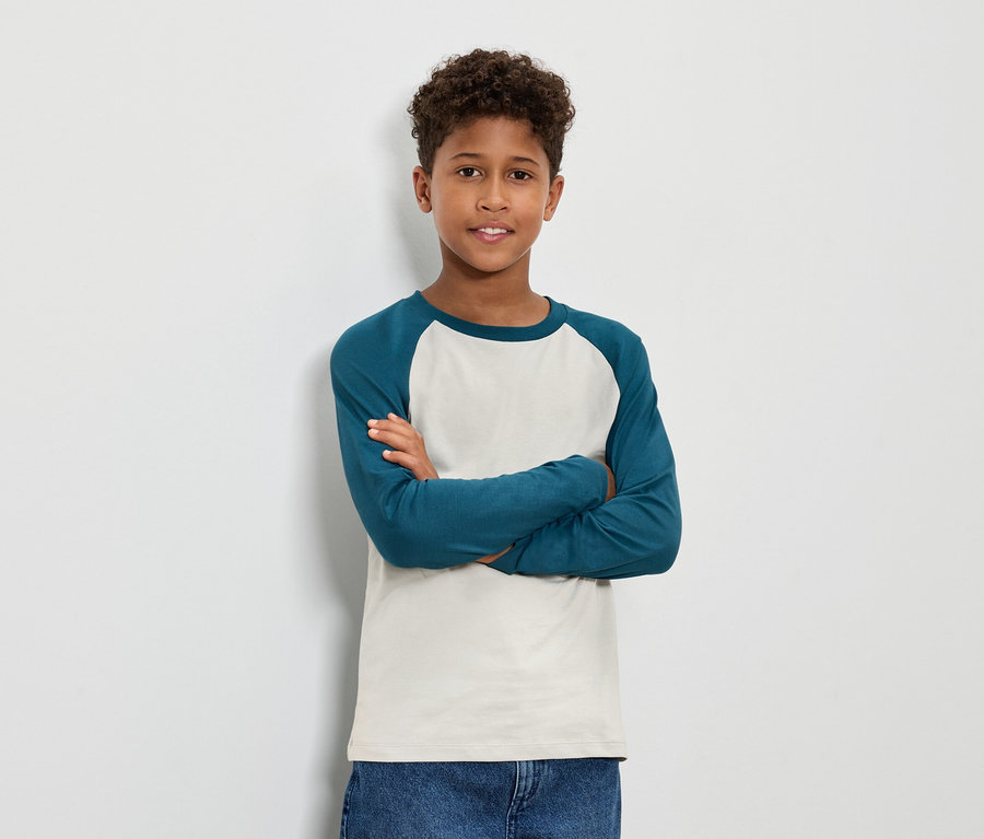 Chlapec s hnedými kučeravými vlasmi stojí s prekríženými rukami a má na sebe modrozelené tričko s dlhým rukávom a džínsy.