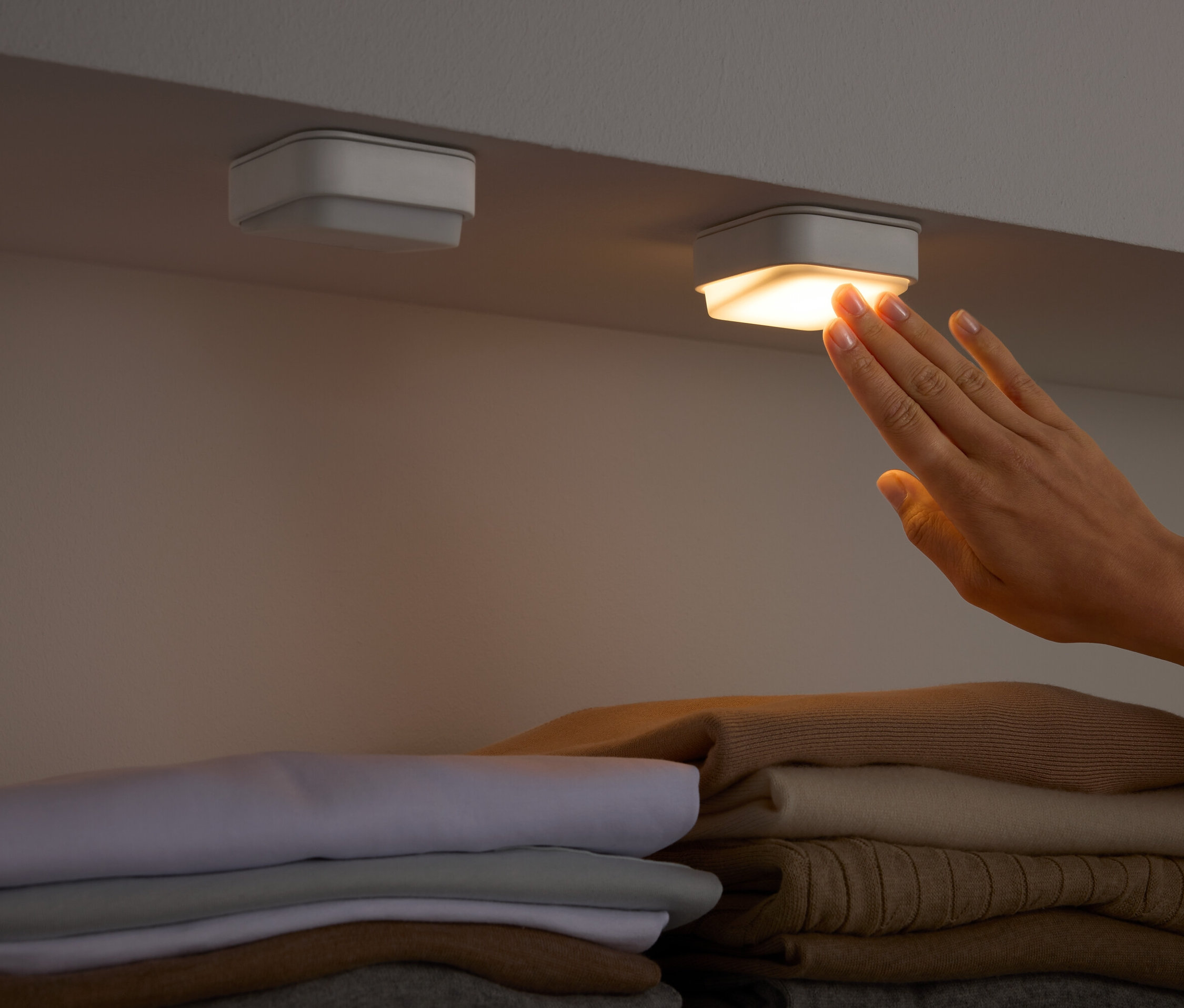 Ruka sa dotýka štvorcového svetla pod poličkou nad zloženým oblečením.