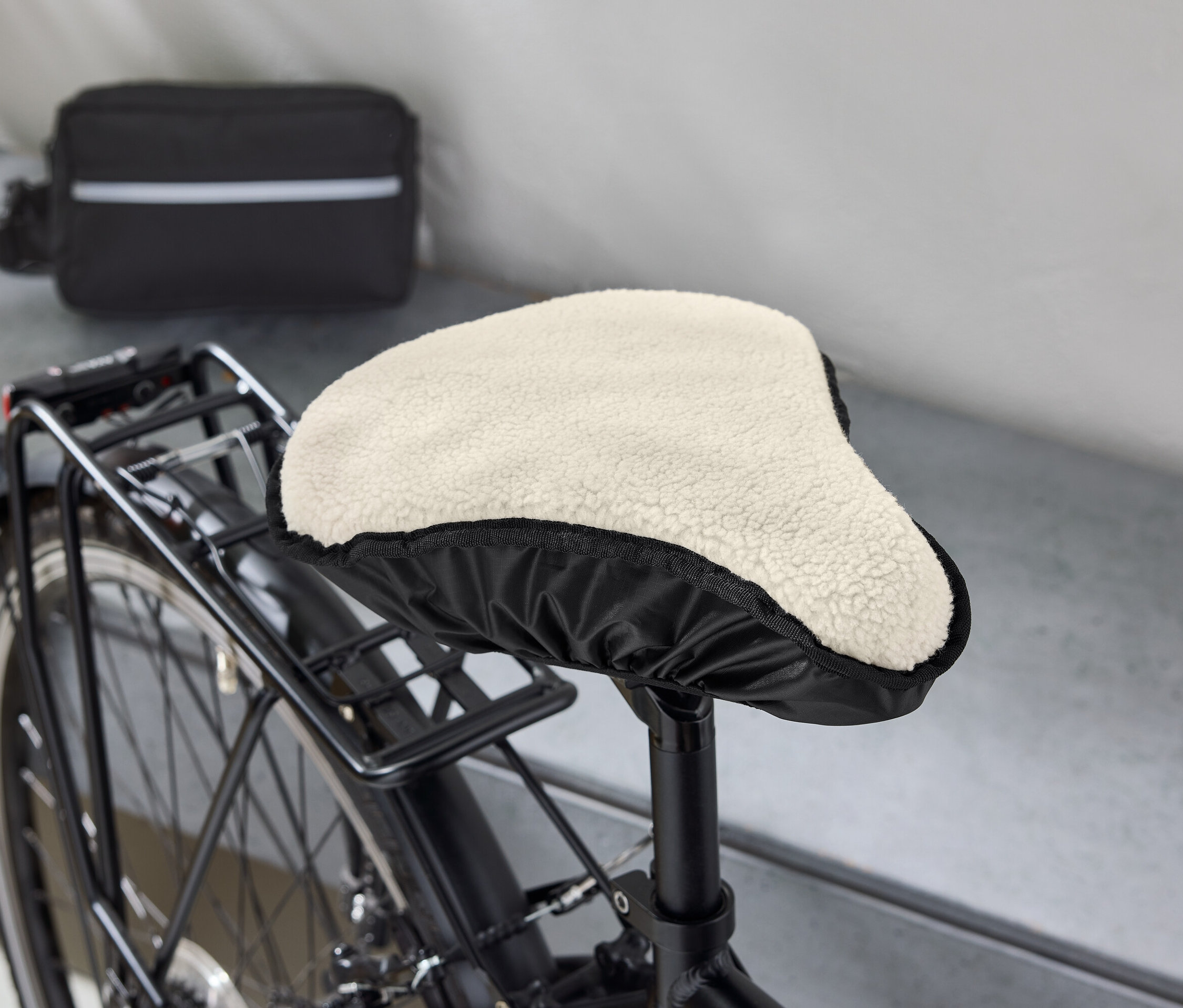 Detail bicykla s čiernym rámom, ktorého sedlo je pokryté béžovým, chlpatým poťahom. V pozadí je vidieť čiernu tašku s bielym pruhom.