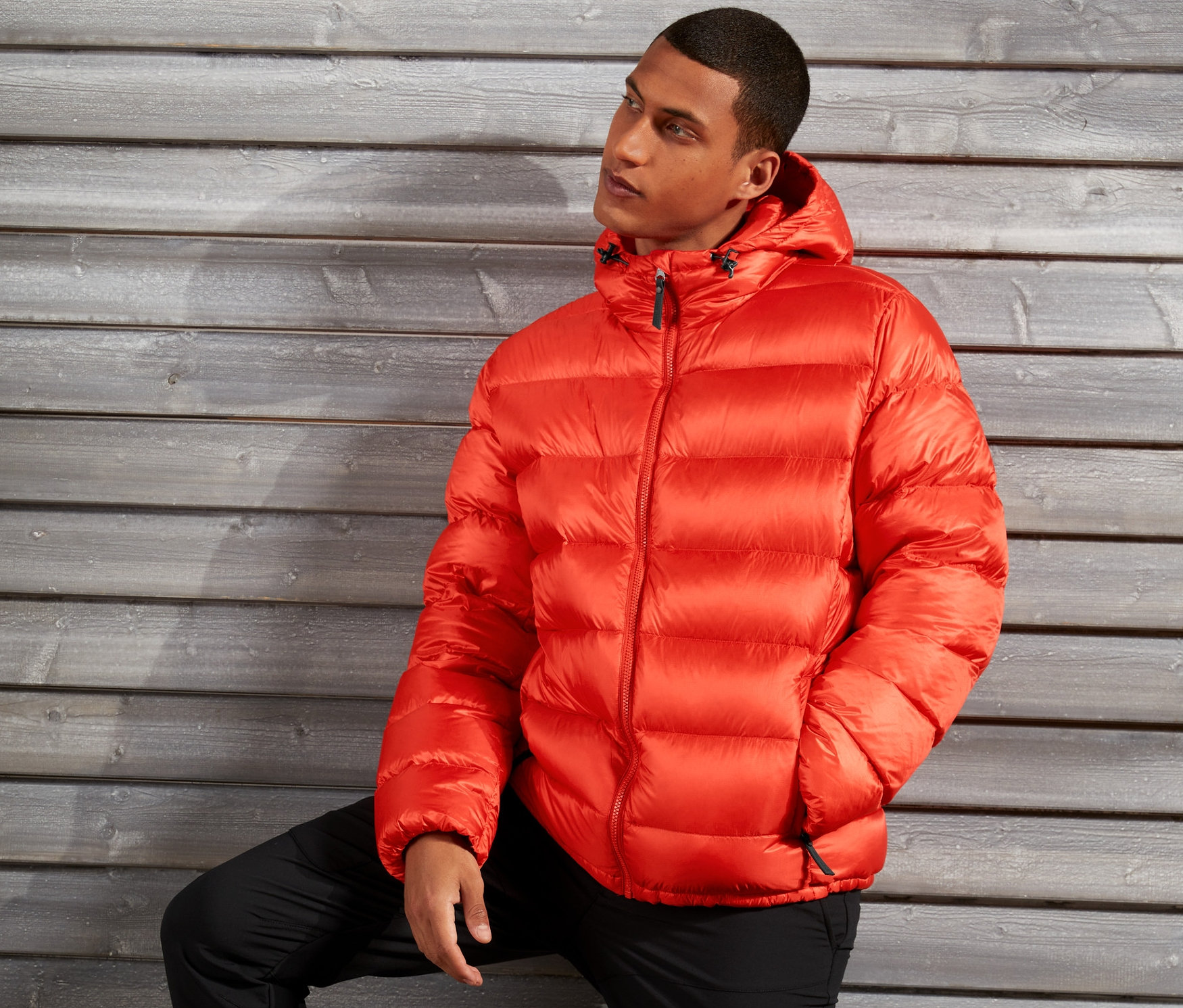 Muž pózuje v oranžovej páperovej bunde pred drevenou stenou.