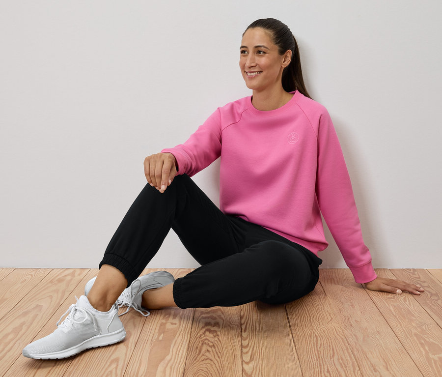 Žena sedí na drevenej podlahe, má na sebe ružovú mikinu, čierne nohavice a biele tenisky.