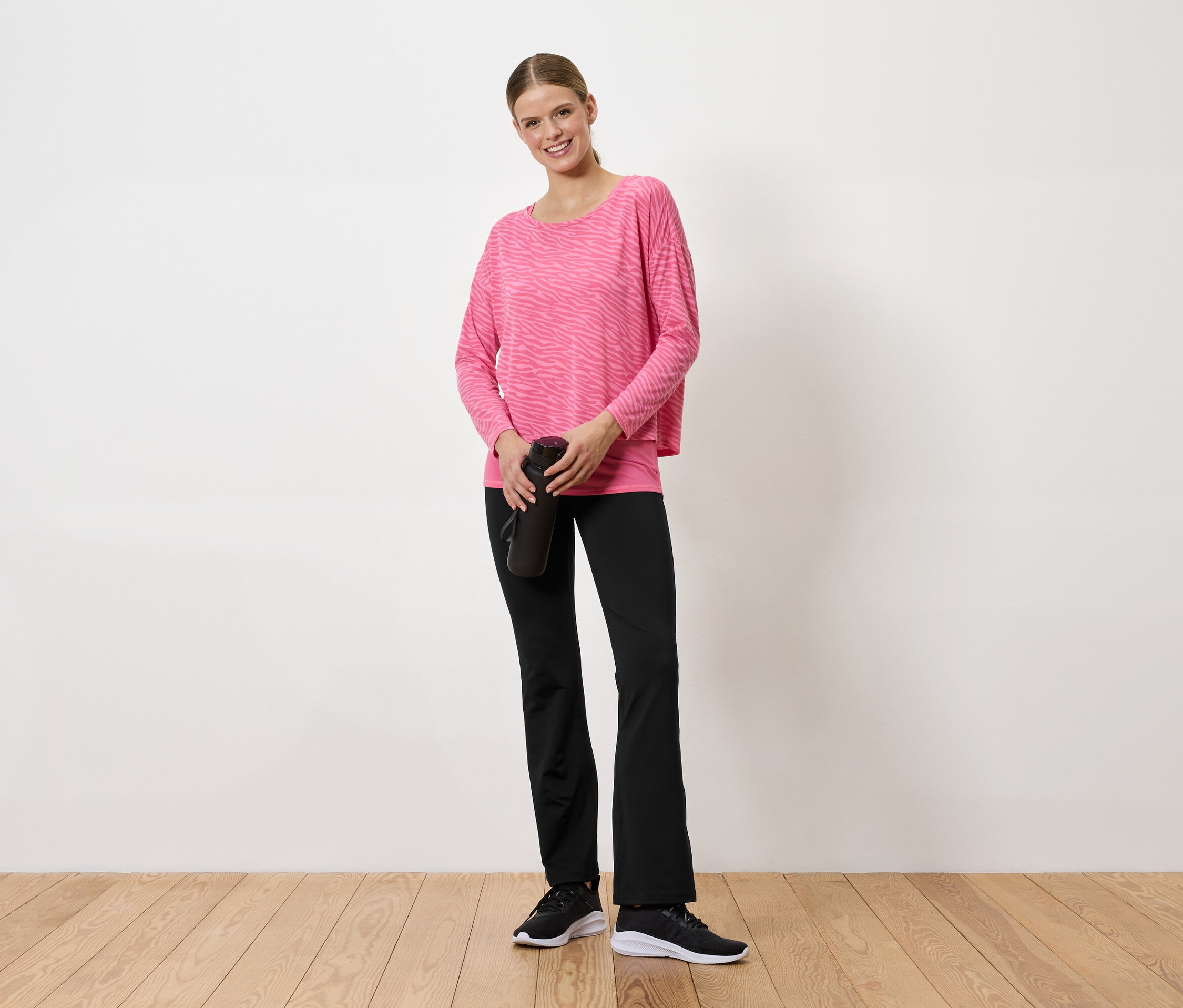 Žena v ružovom svetri a čiernych nohaviciach drží v rukách čiernu fľašu na vodu.