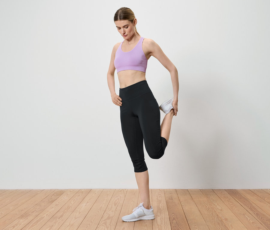 Žena si naťahuje nohu držaním členku v ruke. Má na sebe fialovú športovú podprsenku, čierne legíny a biele tenisky.