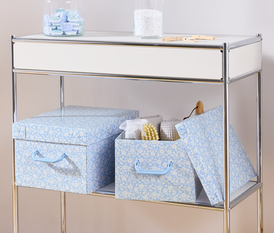 Na polici stoja dva modré úložné boxy s vekom a úložný box s vekom vnútri s bielymi uterákmi a kefou.