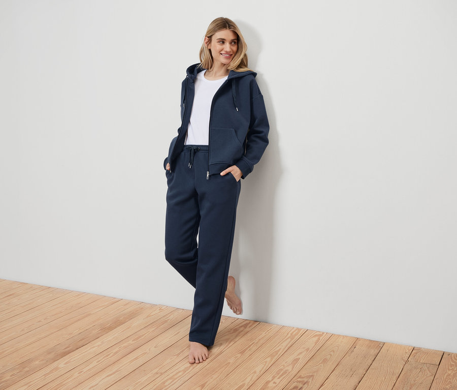 Žena v tmavomodrej teplákovej súprave opretá o bielu stenu.