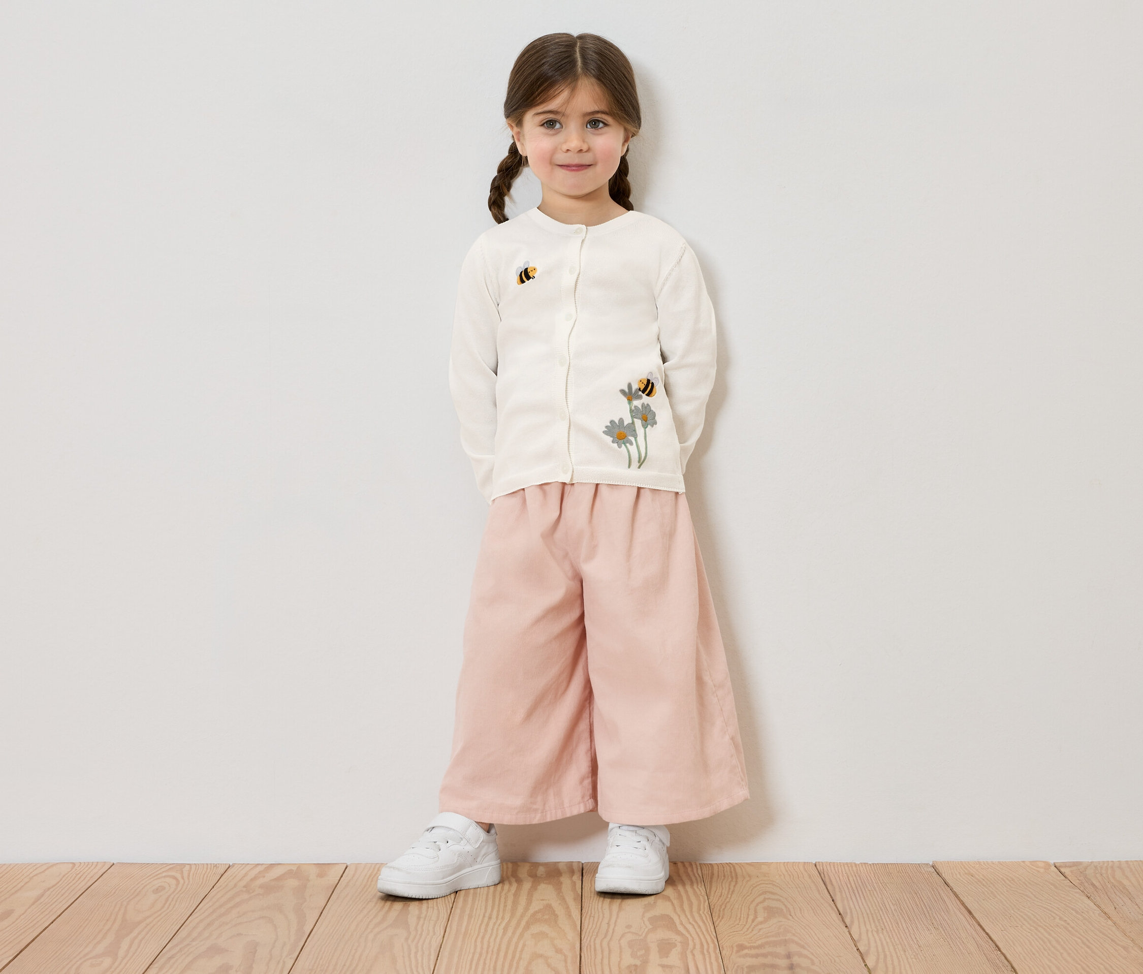 Dievča so zapletanými vlasmi má na sebe krémový kardigán s kvetinovou a včelou výšivkou, ružové nohavice a biele tenisky, na pozadí bielej steny.