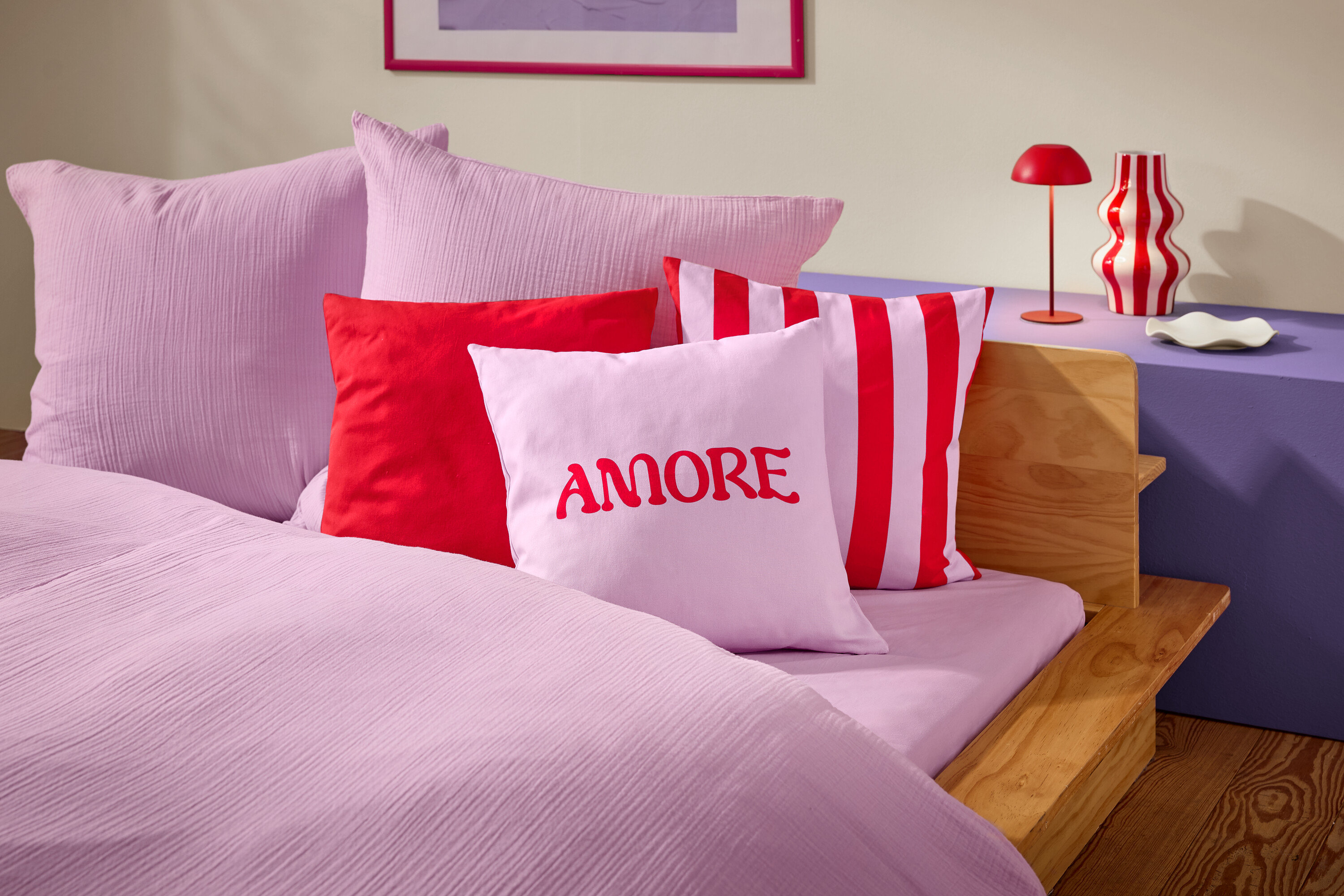 Detailný záber postele s ružovou posteľnou bielizňou a vankúšmi. Na jednom z vankúšov je červeným písmom napísané slovo AMORE.