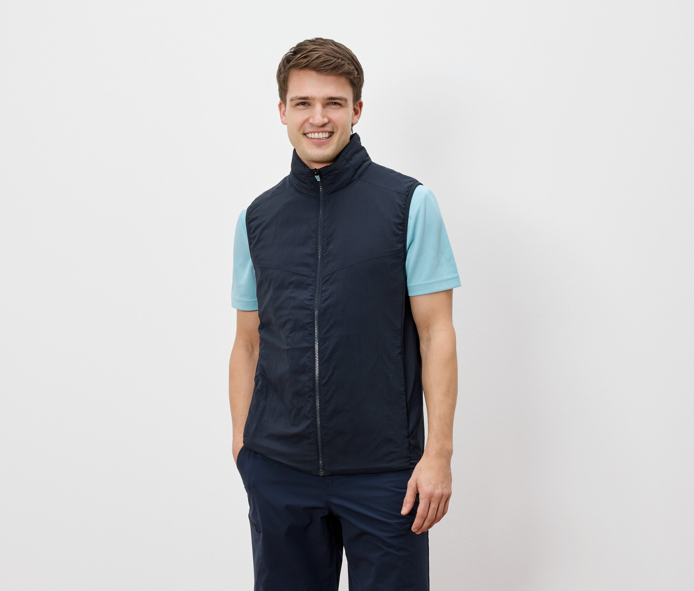 Muž sa usmieva do kamery a má na sebe tmavomodrú vestu cez svetlomodré tričko.
