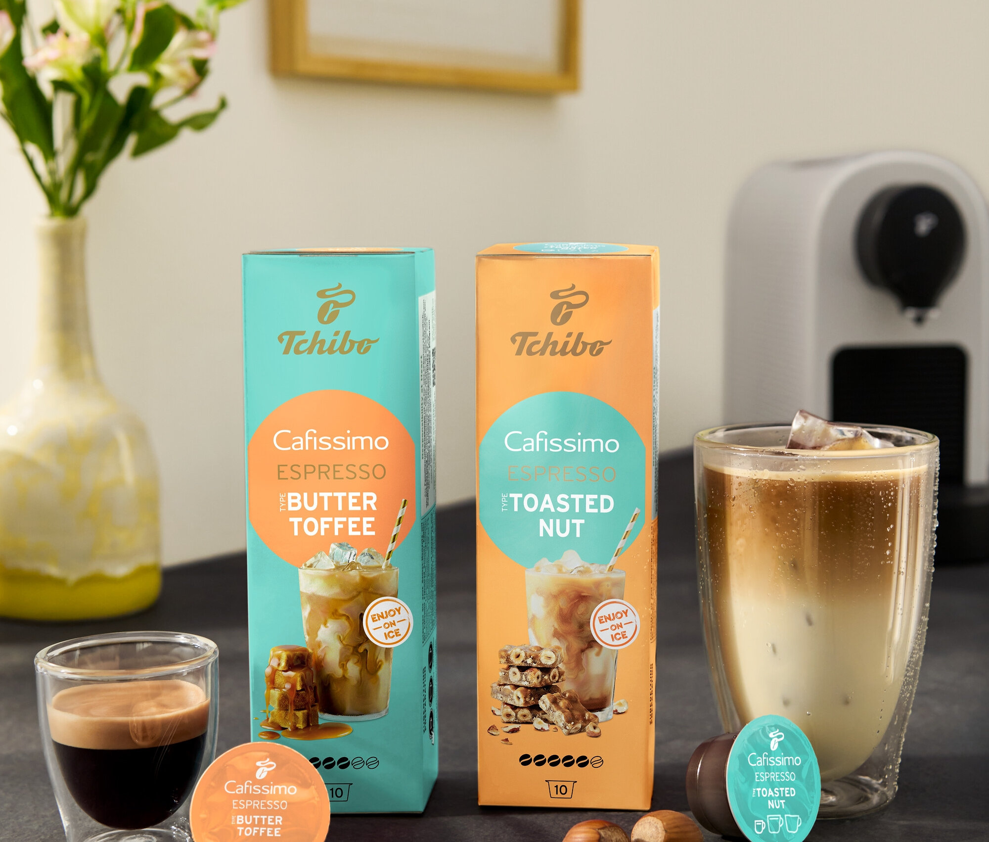 Na kuchynskej linke stojí biely kávovar Cafissimo pure+, šálka espressa, pohár ľadovej kávy, miska s karamelkami a balenia kapsúl Cafissimo Flavoured Espresso – Toasted Nut a Buttertoffee.