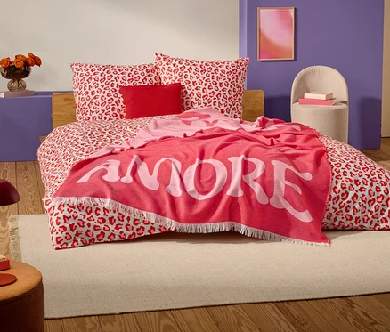 Na posteli s červeno-bielym leopardím vzorom leží červená deka s nápisom "AMORE" veľkými bielymi písmenami.
