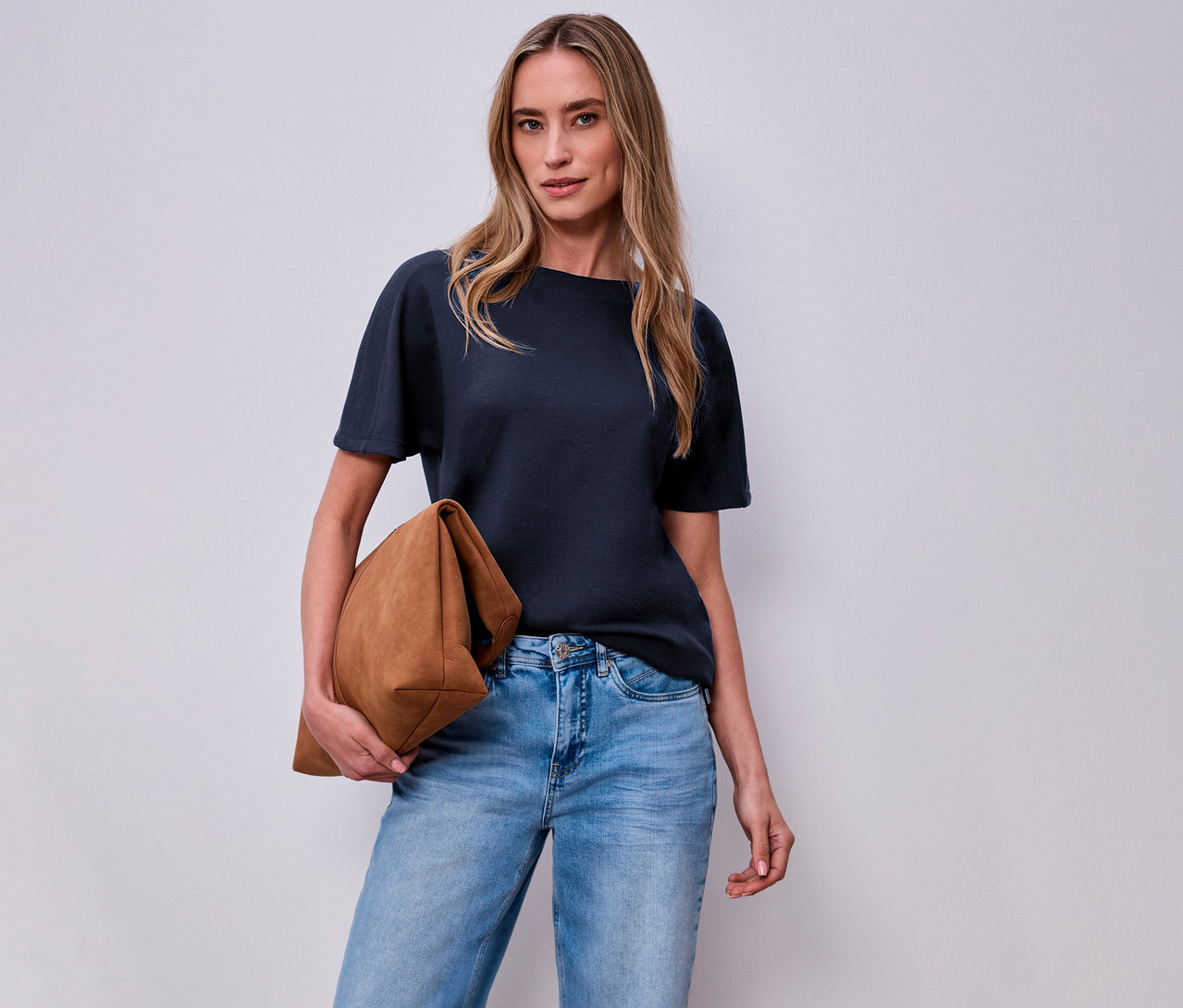Žena s hnedou taškou a námorníckym modrým tričkom s krátkym rukávom Street One pred bielou stenou.