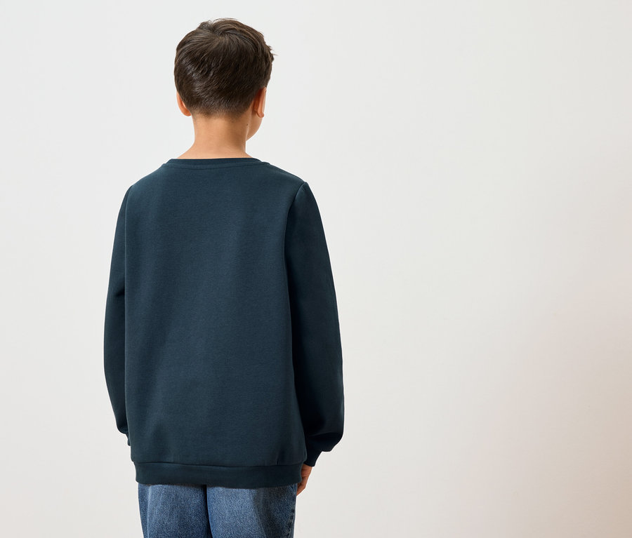 Pohľad zozadu na dieťa s tmavými vlasmi v tmavomodrej mikine a modrých džínsoch na bielom pozadí.