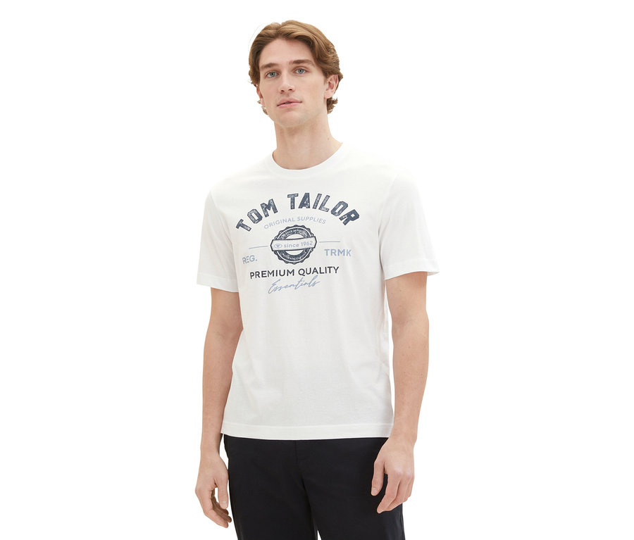 Muž má na sebe biele tričko Tom Tailor s logom a čierne nohavice.