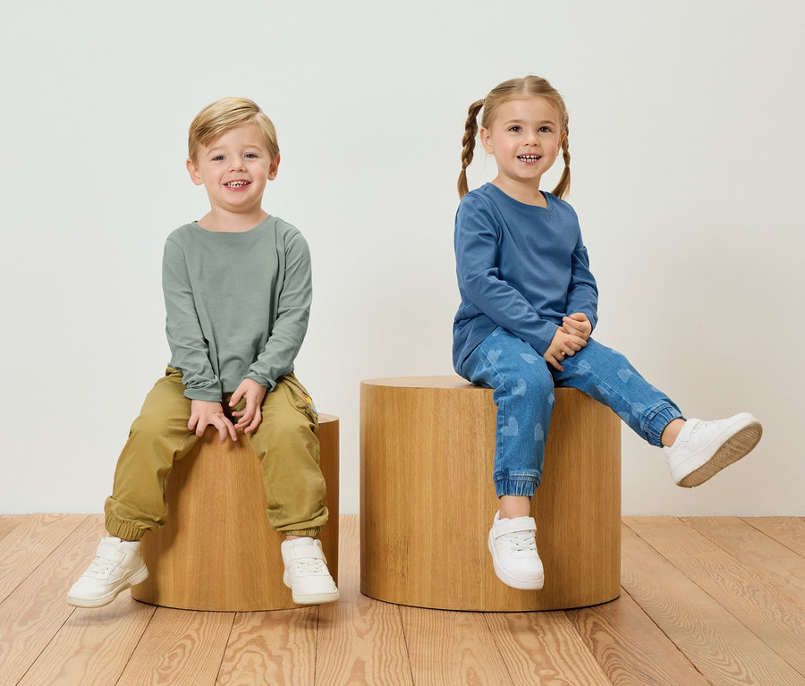 Chlapec v zelenom tričku s dlhým rukávom a khaki nohaviciach sedí vedľa dievčaťa v modrom tričku s dlhým rukávom a džínsoch so vzorom srdca na drevených stoličkách.