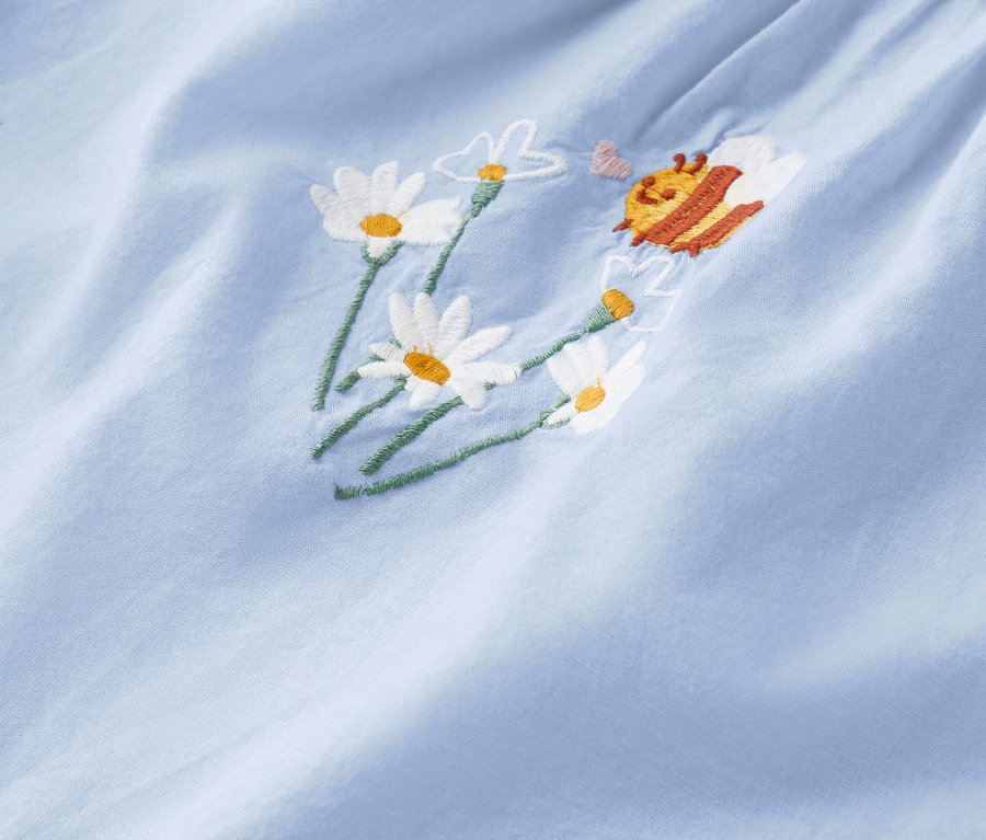 Detailný záber svetlomodrej látky s vyšitými margarétkami a včelou.