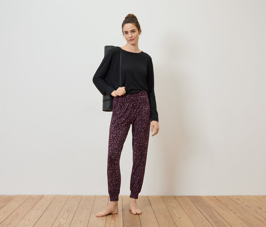 Žena s drdolom má na sebe čierny top, vzorované nohavice a podložku na jogu prehodenú cez rameno, bosá na drevenej podlahe pred bielou stenou.