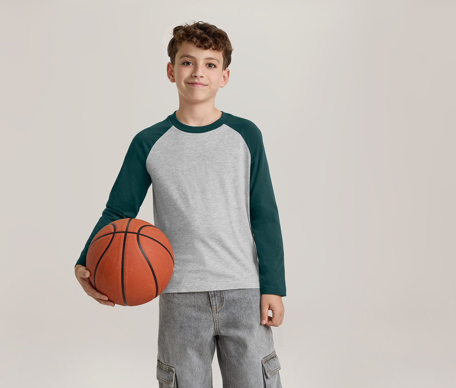 Usmievavý chlapec v sivo-tmavozelenom tričku s dlhým rukávom a sivých cargo nohaviciach drží v ľavej ruke basketbalovú loptu.