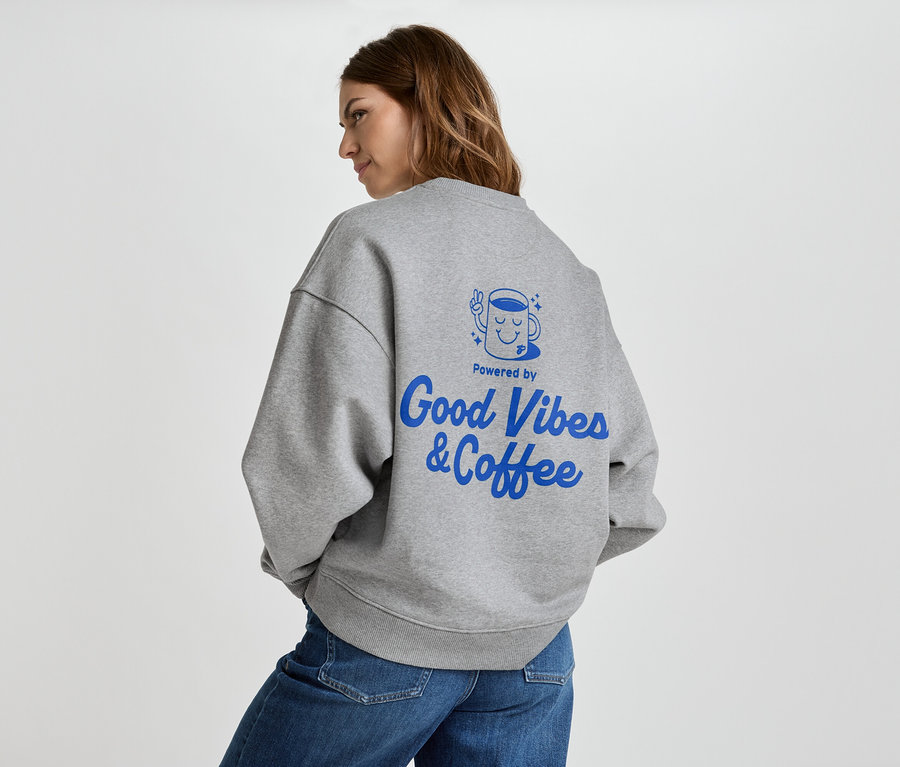 Žena má na sebe sivú mikinu s modrým nápisom "Good Vibes & Coffee" na chrbte a džínsy.