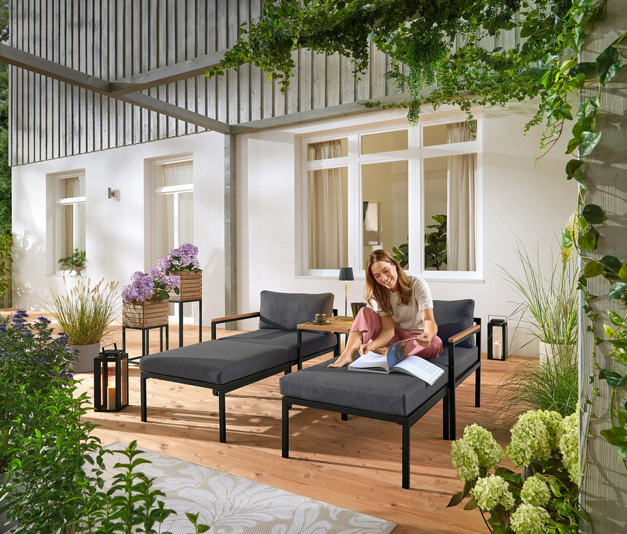 Žena sedí na sivom záhradnom ležadle a číta si knihu. V pozadí sú viditeľné dva stĺpy na rastliny „Elin“.