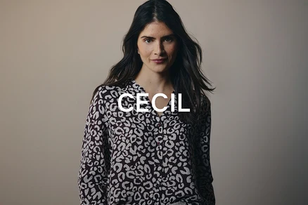 Žena s dlhými tmavými vlasmi, oblečená v tmavohnedej blúzke so svetlým leopardím vzorom, pozerá priamo do kamery.