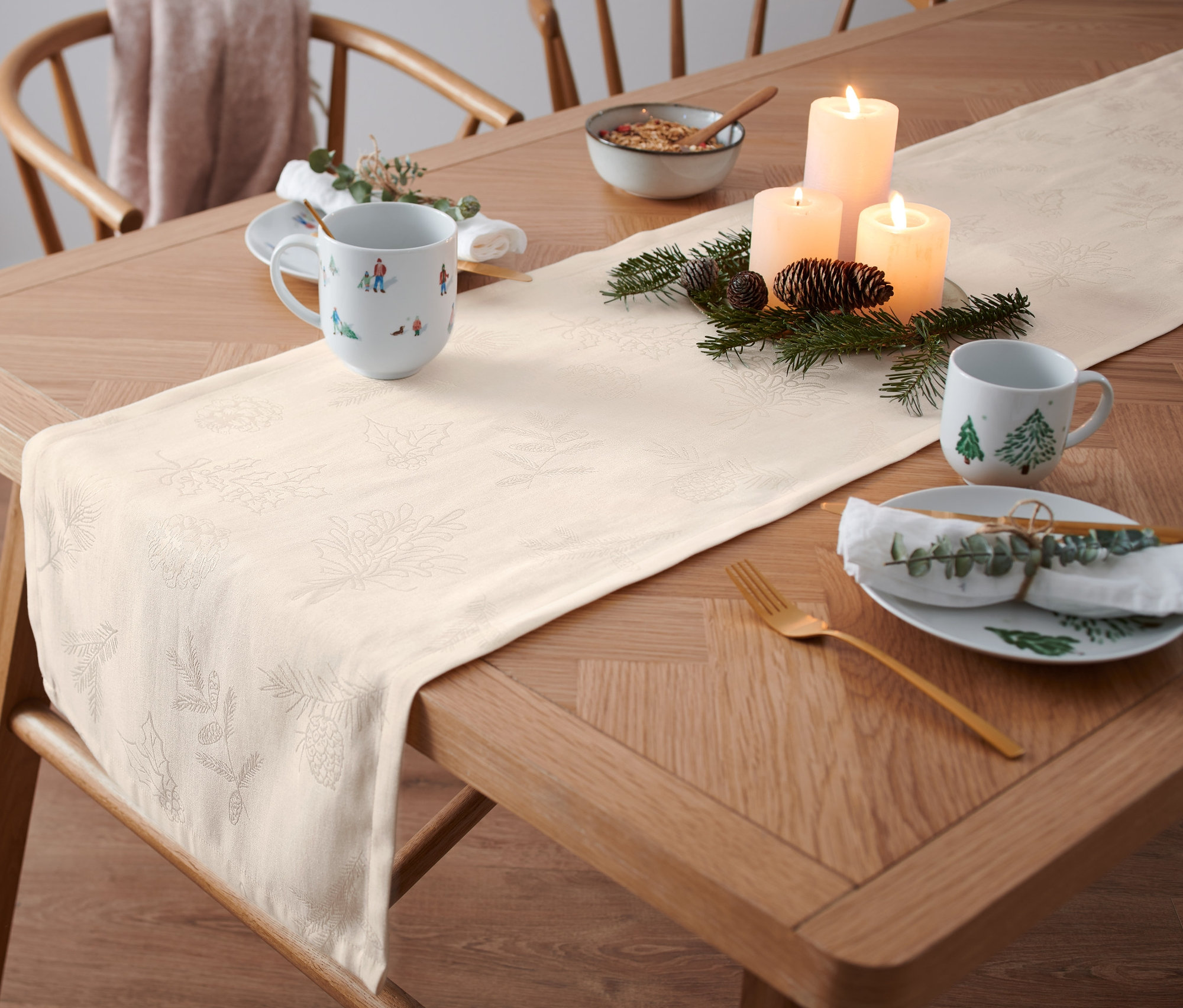 Na drevenom stole leží béžový žakárový behúň. Stôl je prestretý s taniermi, hrnčekmi a sviečkami.