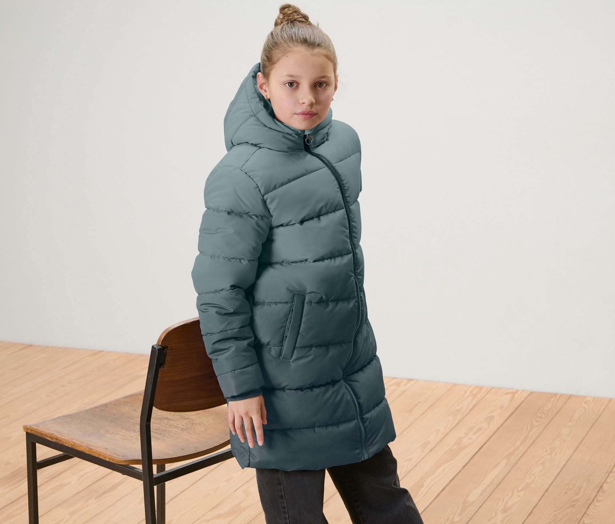 Dievča má na sebe detský termokabát s farebným prechodom a stojí vedľa drevenej stoličky.