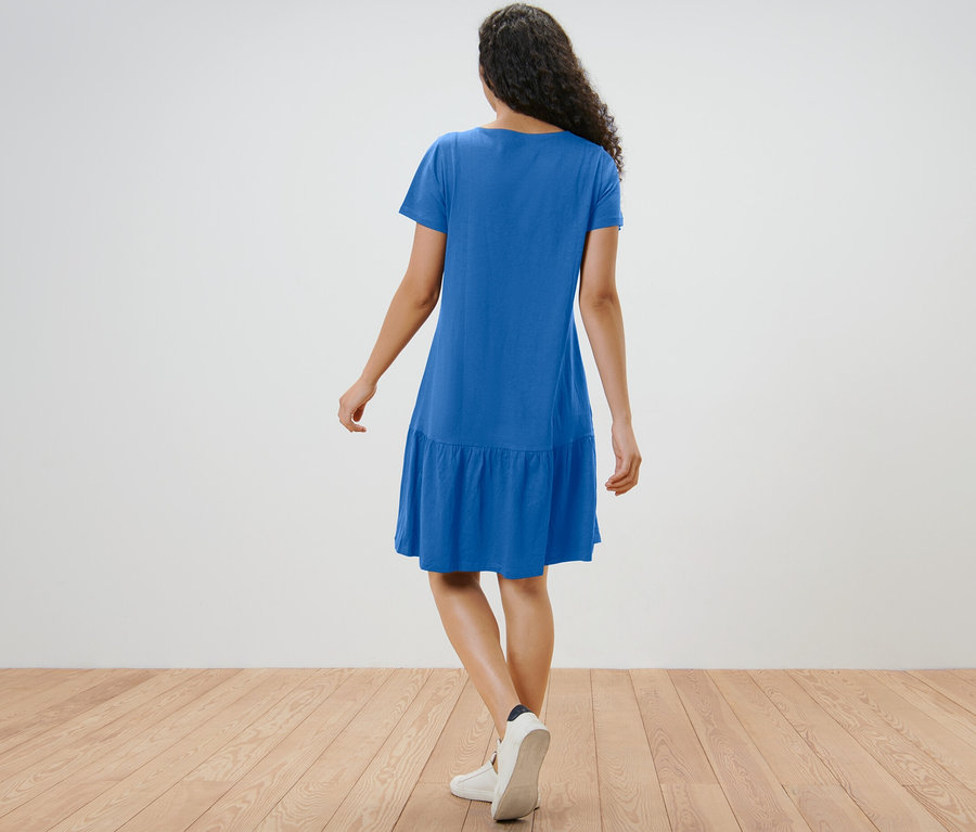 Žena v kobaltovomodrých džersejových šatách s volánom stojí chrbtom k fotoaparátu.