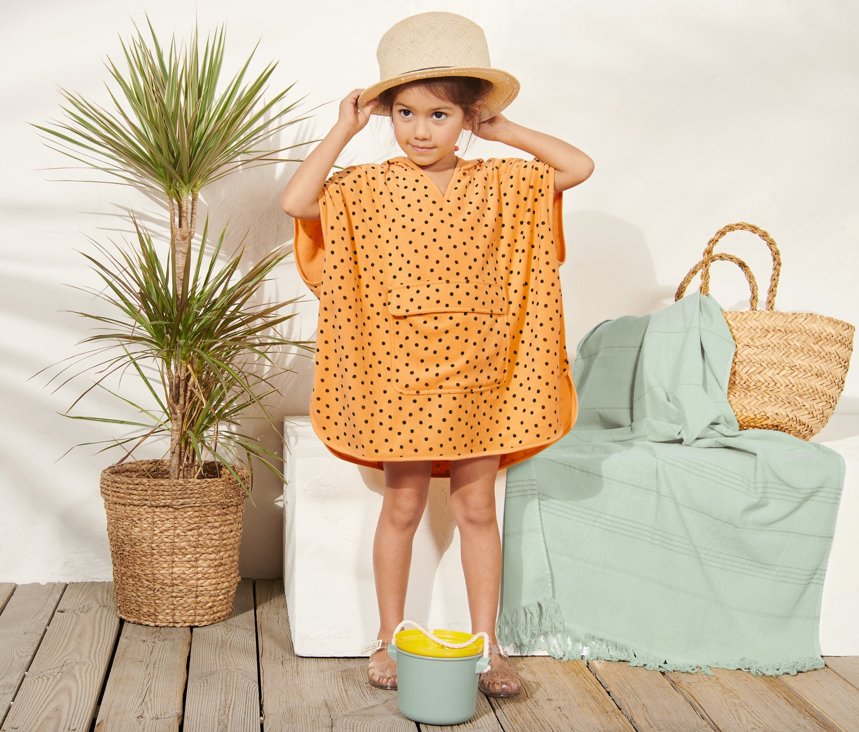 Dievča v oranžovom froté pončo s čiernymi bodkami a slamenom klobúku stojí vedľa rastliny v košíku a plážovej tašky.