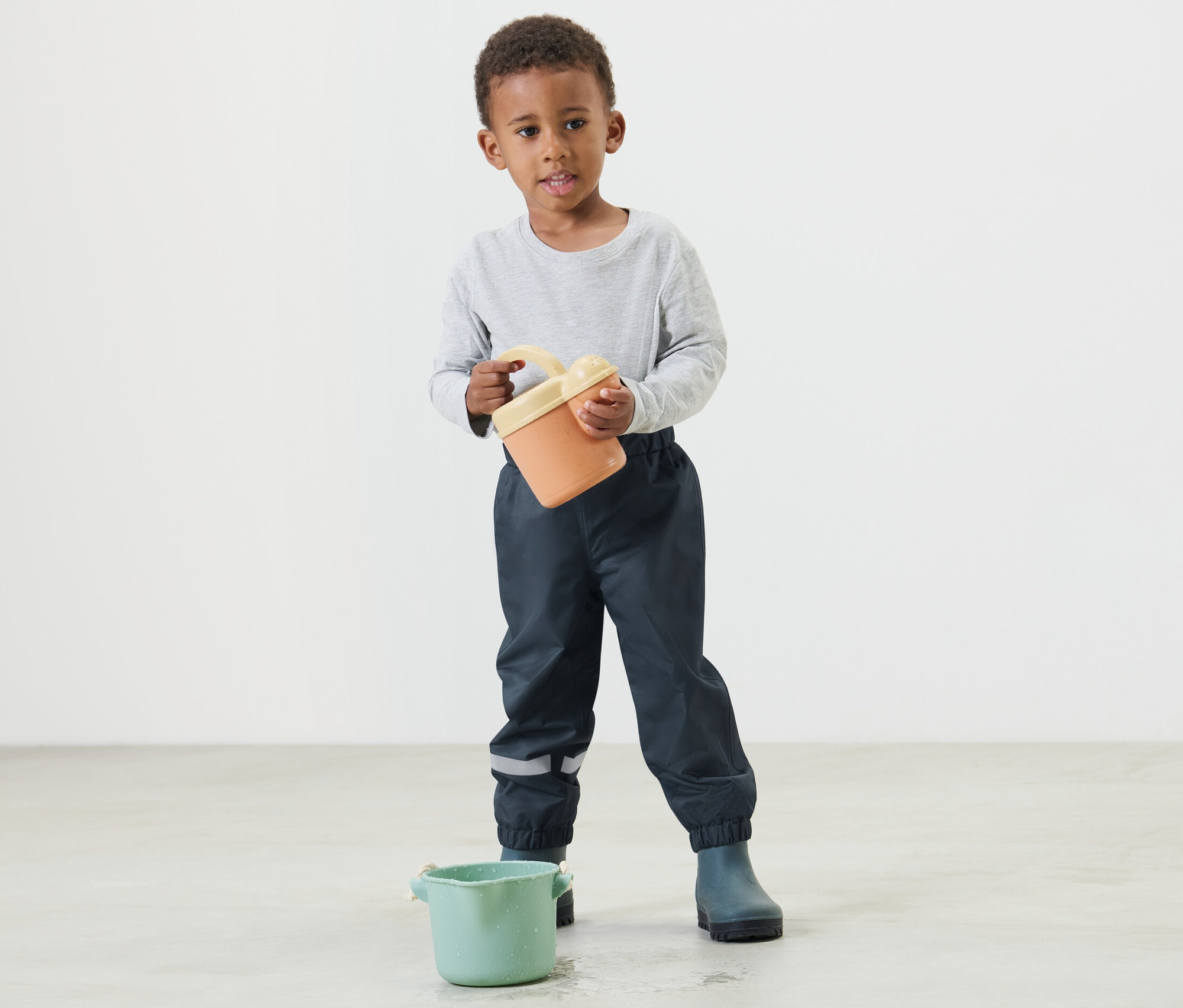 Chlapec drží oranžové vedro a má na sebe tmavomodré detské nohavice do dažďa.