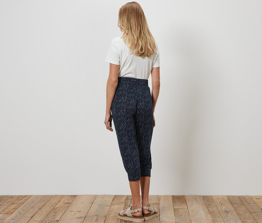 Žena zozadu stojí na drevenej podlahe a má na sebe 3/4 džersejové nohavice modro-krémovobielej farby a biele tričko.