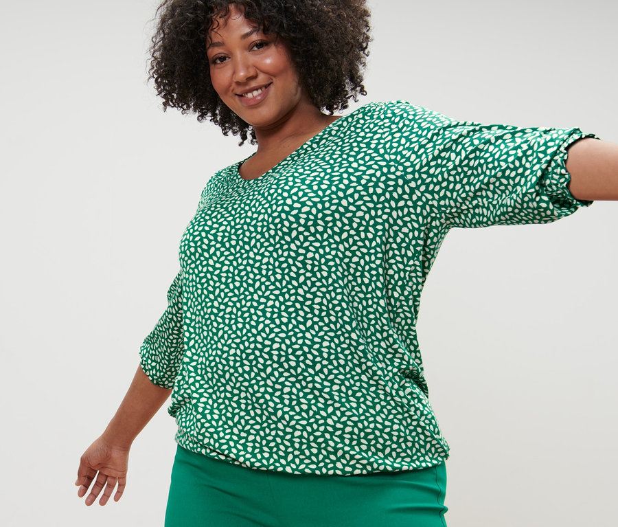 Žena pózuje v zelenom blúzkovom tričku s potlačou a zelených elastických nohaviciach.