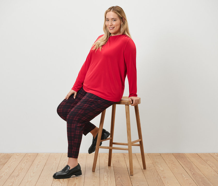 Žena sedí na stoličke a má na sebe kockované elastické nohavice a blúzkové tričko s nariasením.