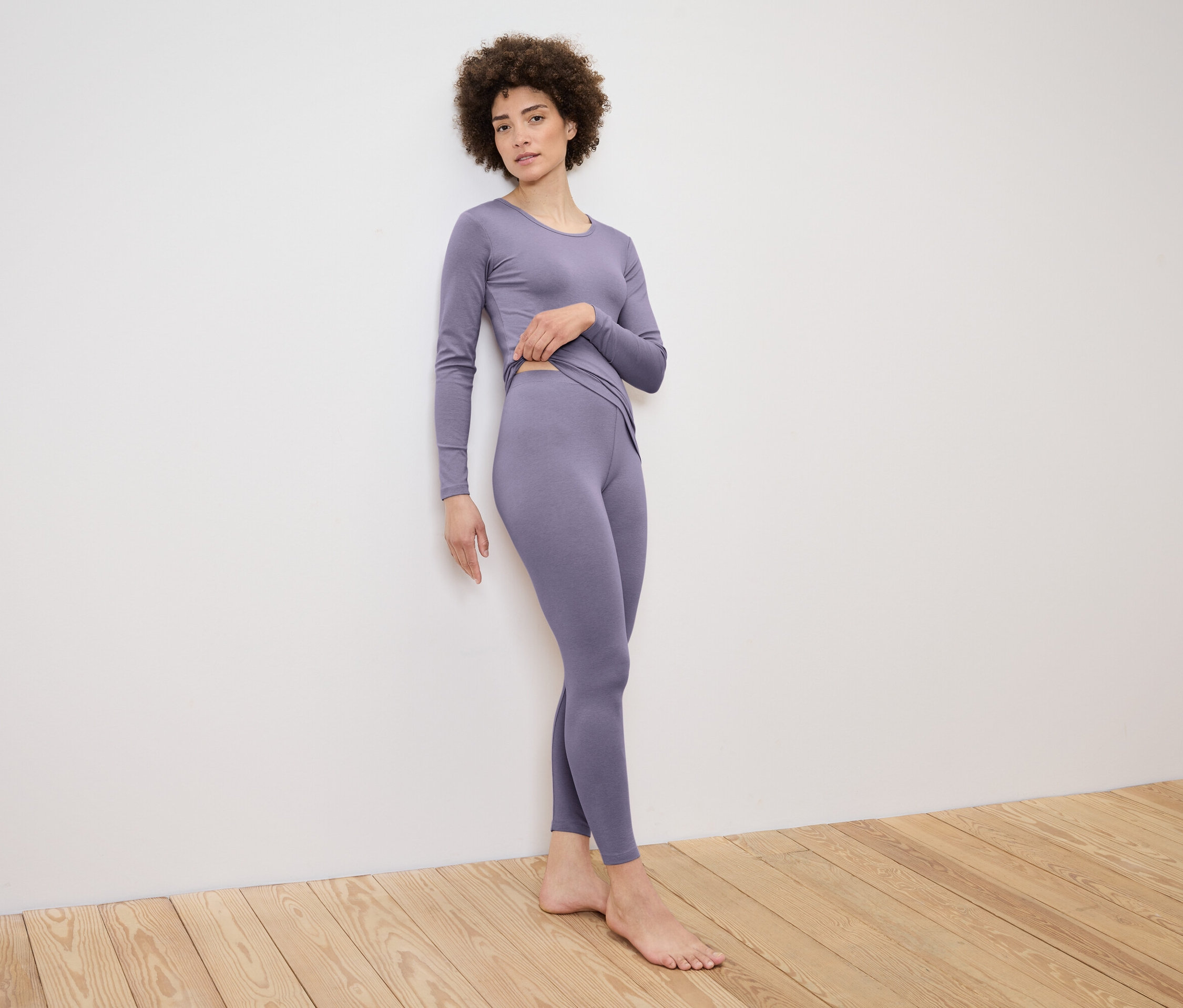 Žena pózuje pri stene v súprave topu s dlhým rukávom a legín vo farbe lila.