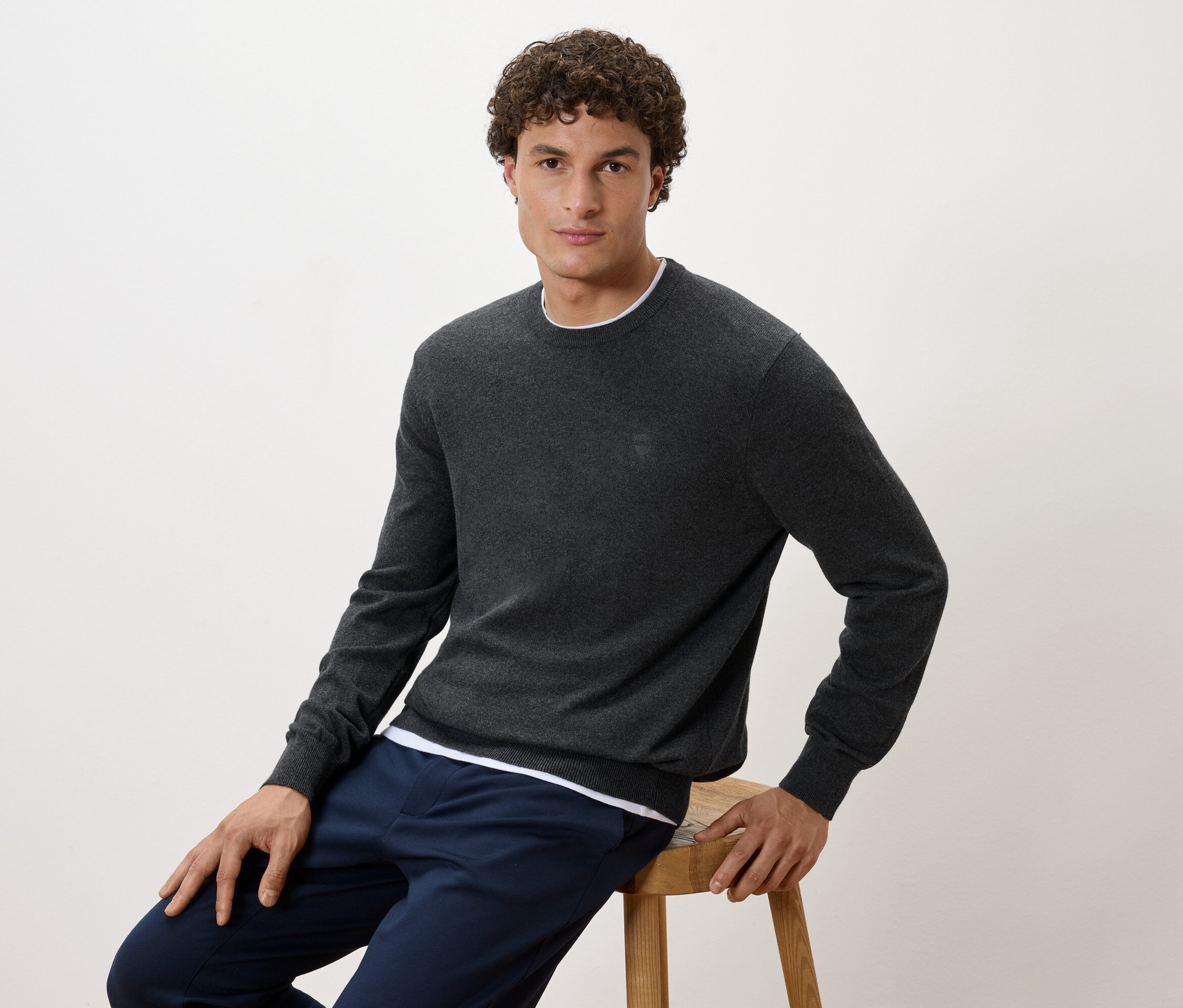Muž sedí na stoličke, oblečený v sivom svetri a tmavomodrých nohaviciach.