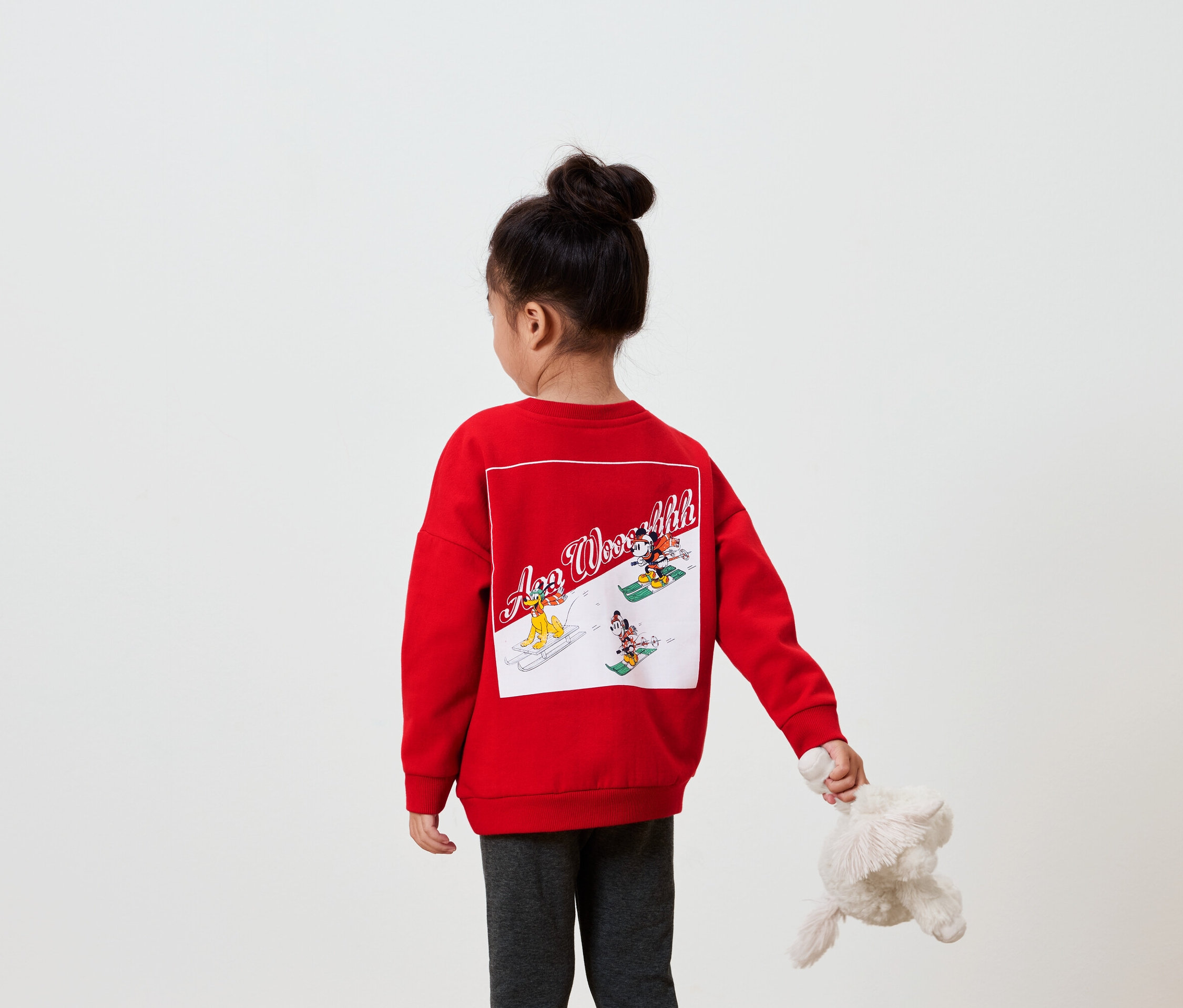 Dievča chrbtom k fotoaparátu má na sebe červenú mikinu s obrázkom na chrbte a v ruke drží plyšovú hračku.