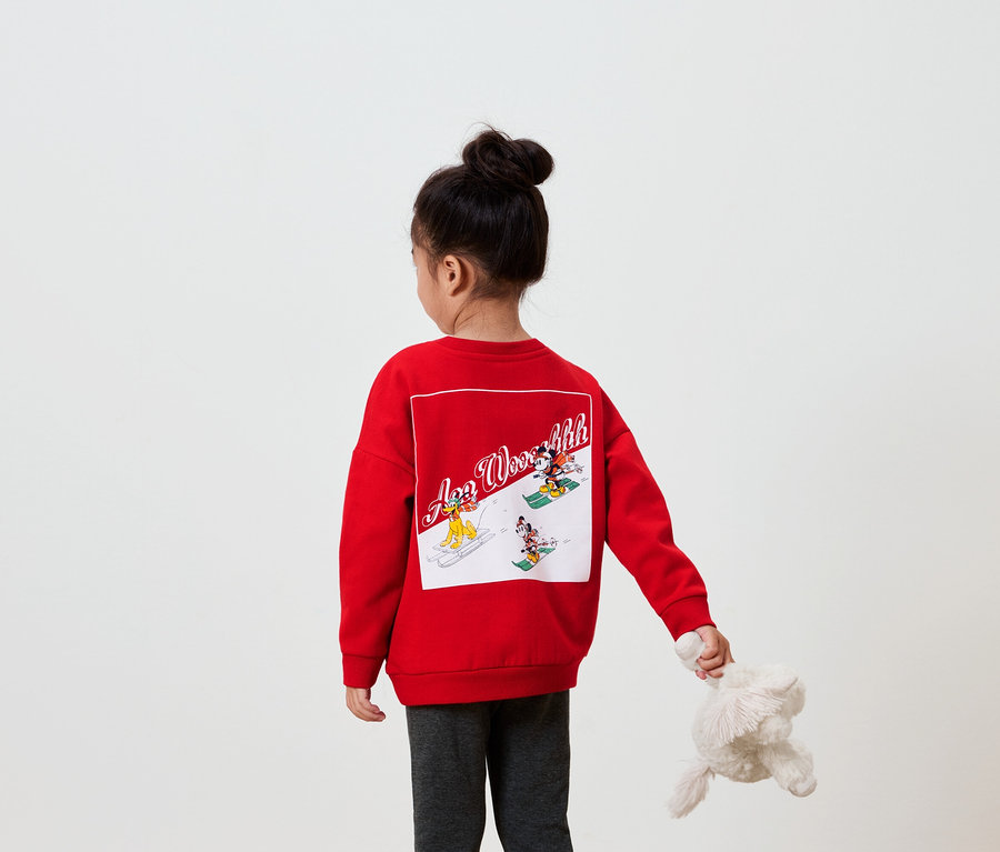 Dievča chrbtom k fotoaparátu má na sebe červenú mikinu s obrázkom na chrbte a v ruke drží plyšovú hračku.