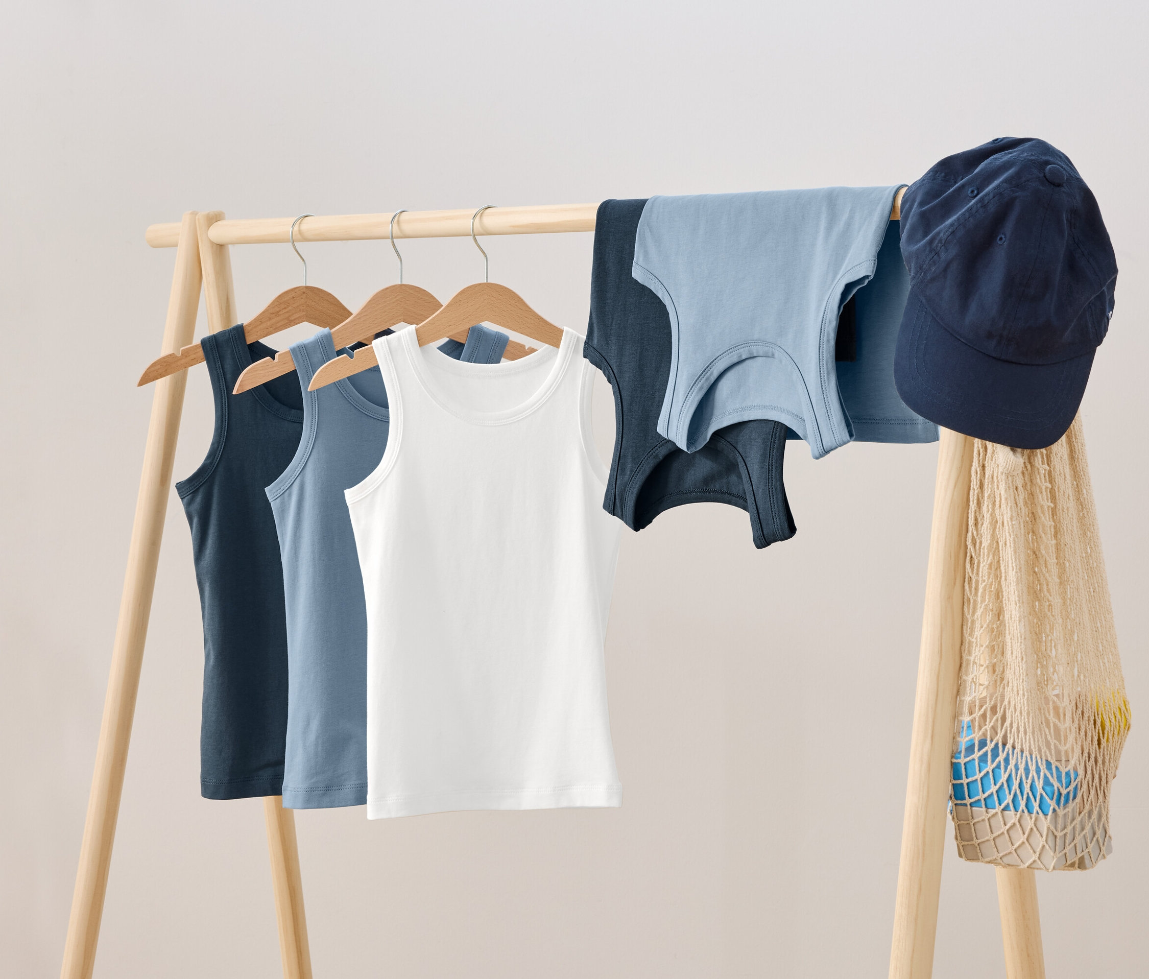 Na drevenom vešiaku visia tričká na vešiakoch, vedľa nich ležia dve ďalšie tričká, modrá šiltovka a sieťovaná taška.