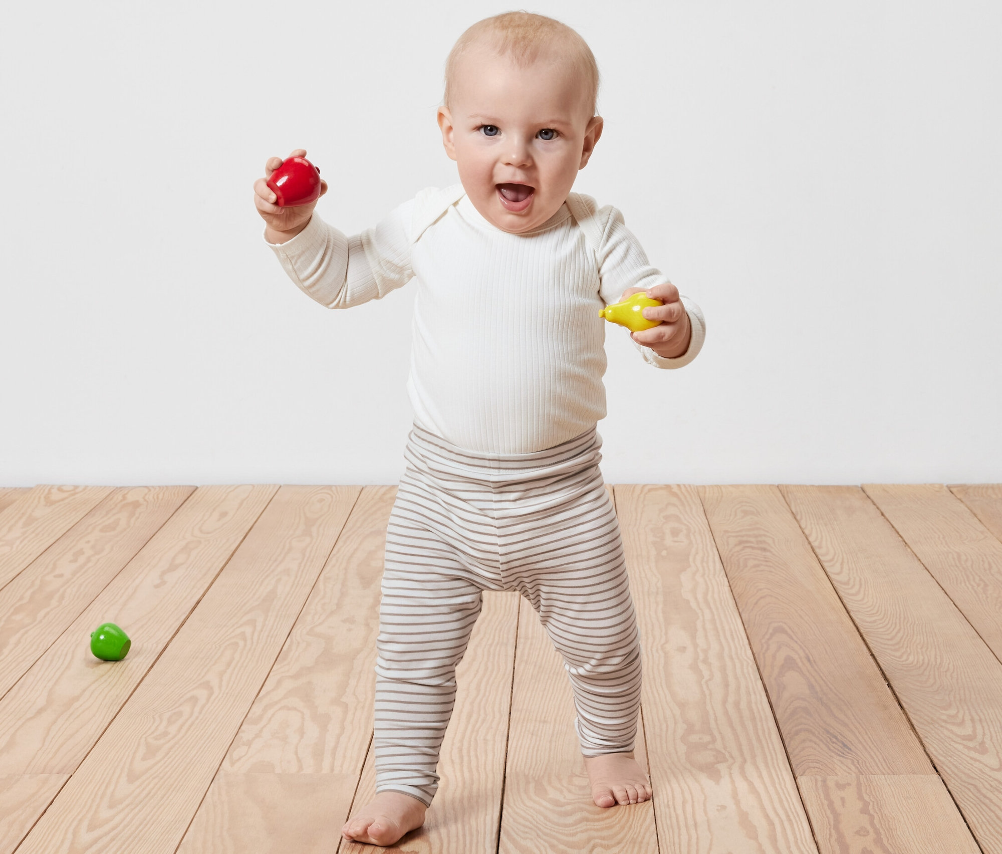 Usmievajúce sa dieťa stojí na drevenej podlahe a drží v rukách červenú a žltú hračku. Oblečené má Detské legíny, 5 ks.