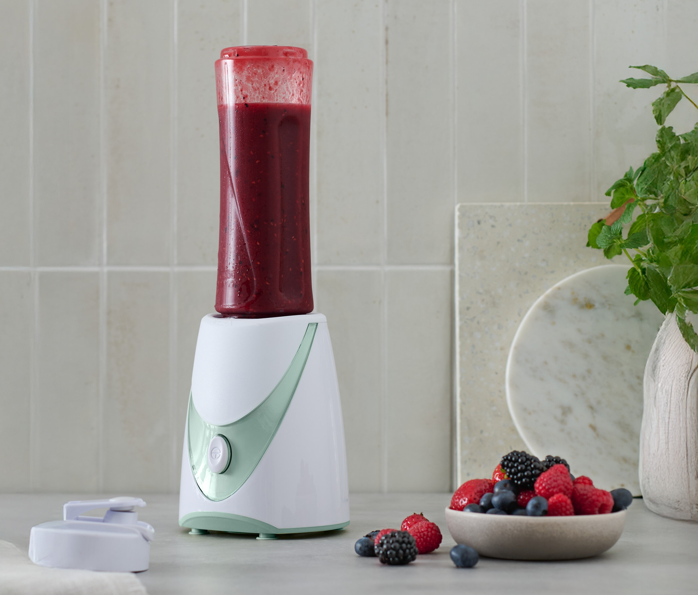 Mini-smoothie maker s hotovým smoothie na vrchu, vedľa miska s bobuľami.
