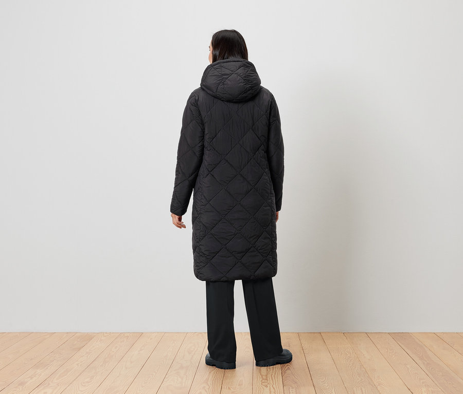 Žena stojí chrbtom k fotoaparátu a má na sebe čierny prešívaný kabát s kapucňou.