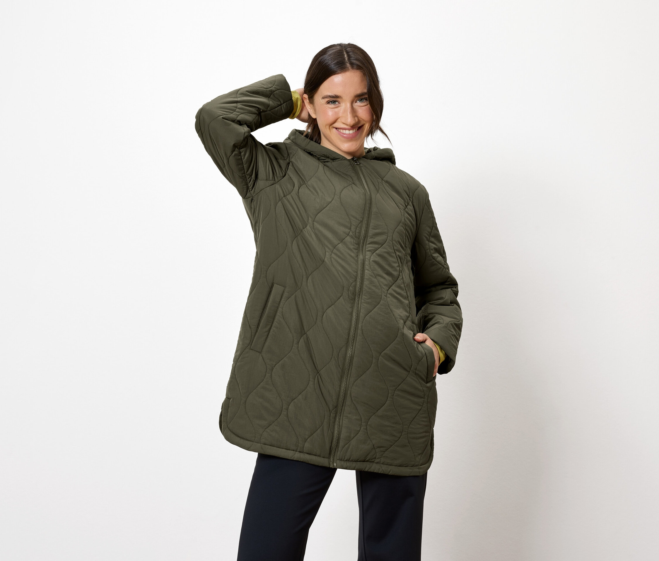 Žena pózuje v olivovozelenej prešívanej bunde s kapucňou a tmavomodrých nohaviciach.